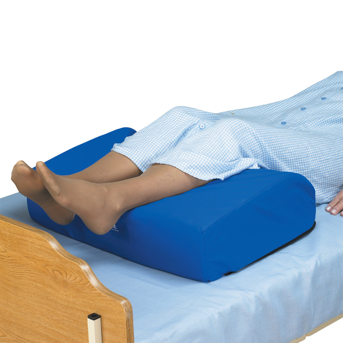 Person's legs on SkiL-Care Heels-Off cushion, elevating heels while lying on a bed with a blue cover and wooden headboard.