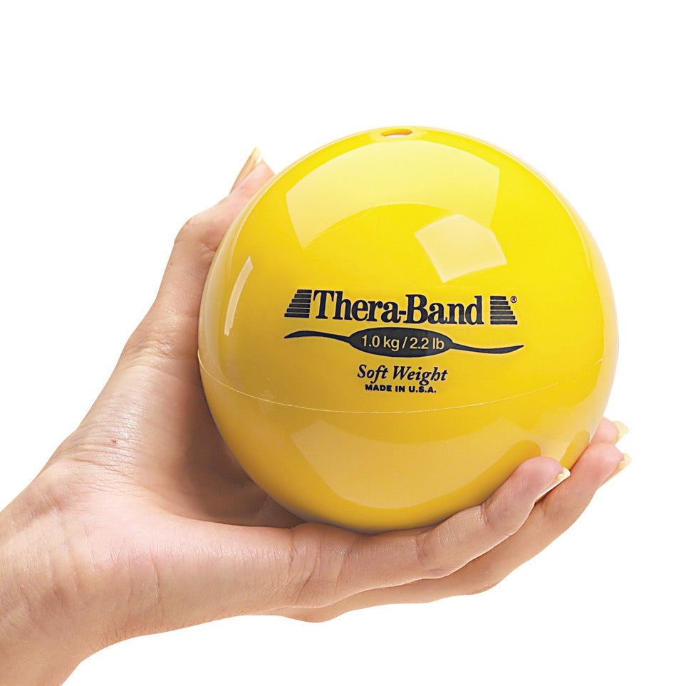 A hand holds a yellow THERABAND Soft Weight ball, labeled 1.0 kg/2.2 lb, made in the USA.
