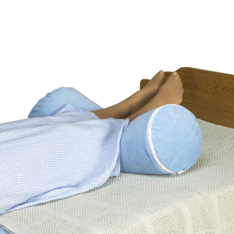 A person rests on a bed with feet elevated by a SkiL-Care Ultra-Soft Bed Bolster. The bolster is light blue and cylindrical.