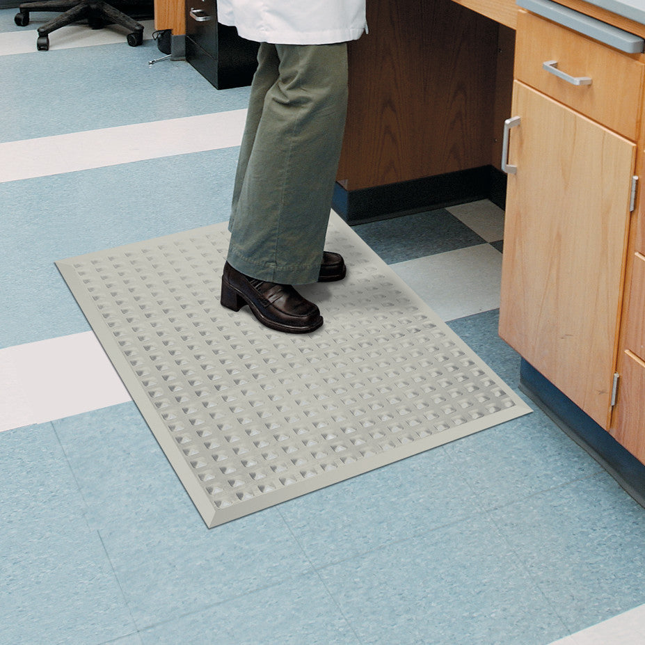 Person standing on a Wearwell Autoclavable Anti-Fatigue Mat in a lab, near wooden cabinetry, on a tiled floor.