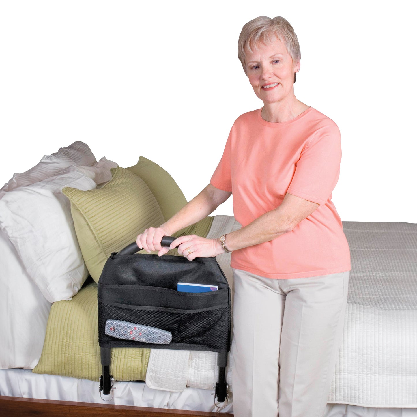 An older woman uses the Bed Rail Advantage Traveler, a black bed rail with a pouch, installed next to a bed with green pillows.