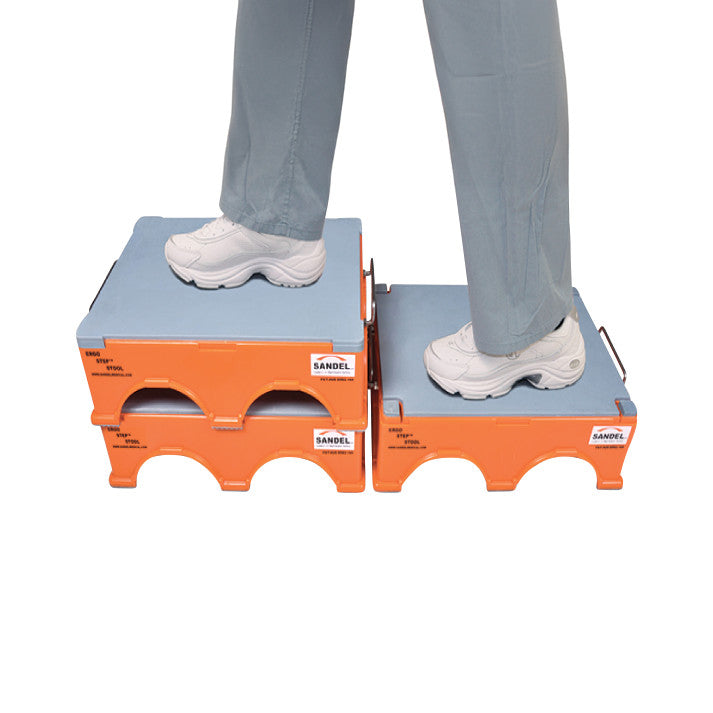 Person standing on stacked orange SANDEL Ergo-Step Stools, wearing white sneakers and gray pants, shown from the knees down.