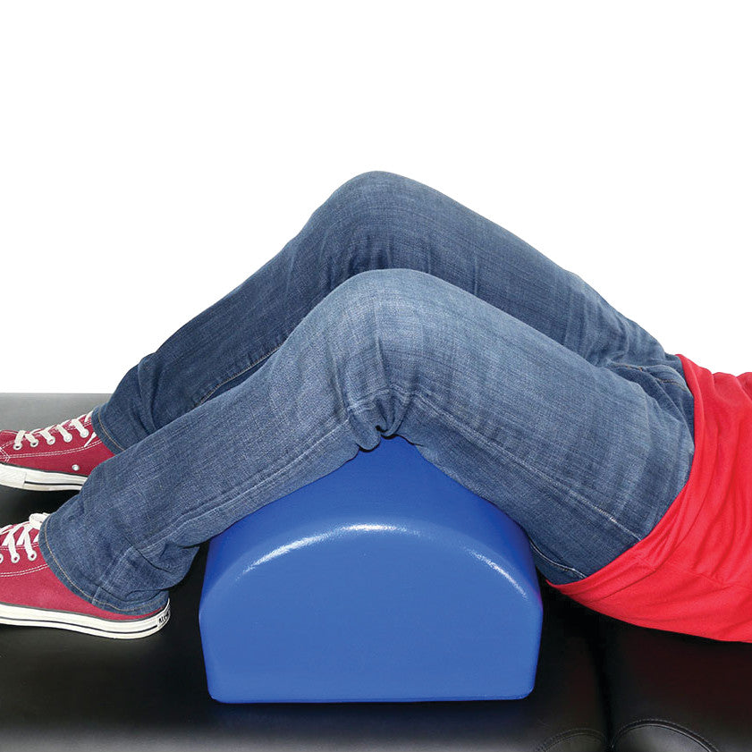 A person uses the Skillbuilders Crescent Knee Bolster, with legs positioned on the blue bolster while lying on a black surface.