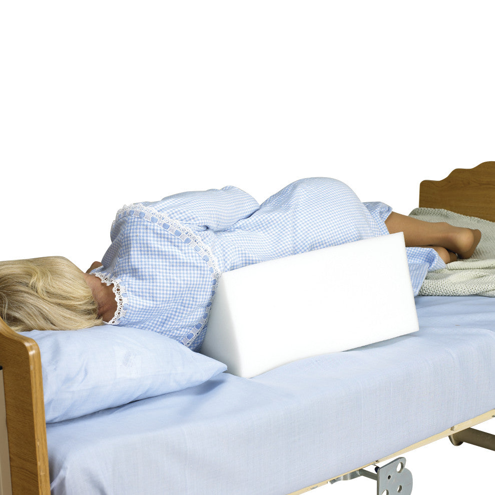 A person lies in bed with a SkiL-Care Positioning Wedge without Cover against their back, on blue sheets.