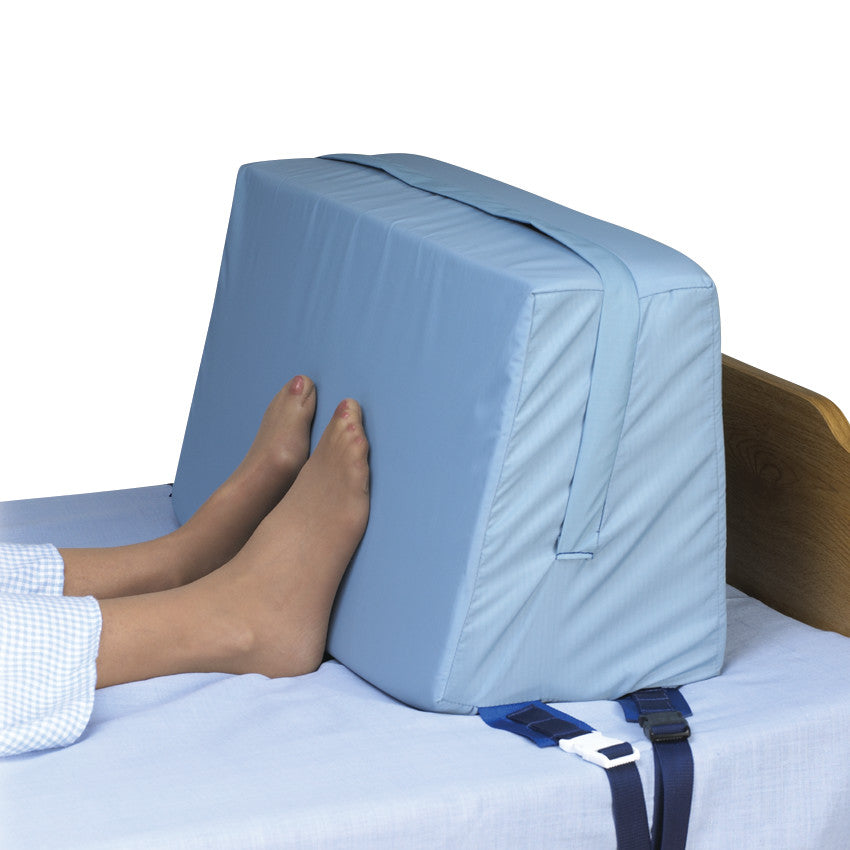 A person rests their feet against the SkiL-Care Bed Foot Support, a blue cushioned block strapped to a bed for support.
