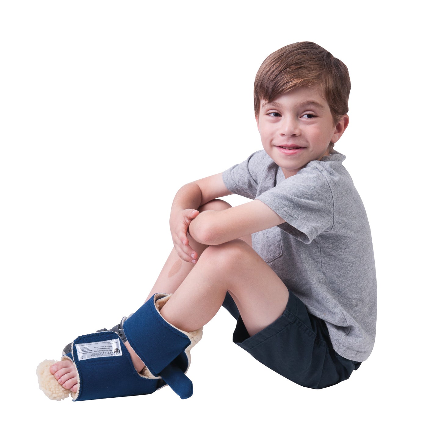 A child wearing a Comfy Splints Pediatric Ambulating Contracture Boot on the foot, sitting cross-legged, smiling, in a grey shirt.