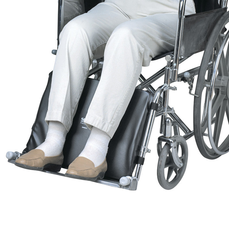 A person in a wheelchair uses the SkiL-Care Wheelchair Leg Pad, which is black and positioned on the footrests for comfort.