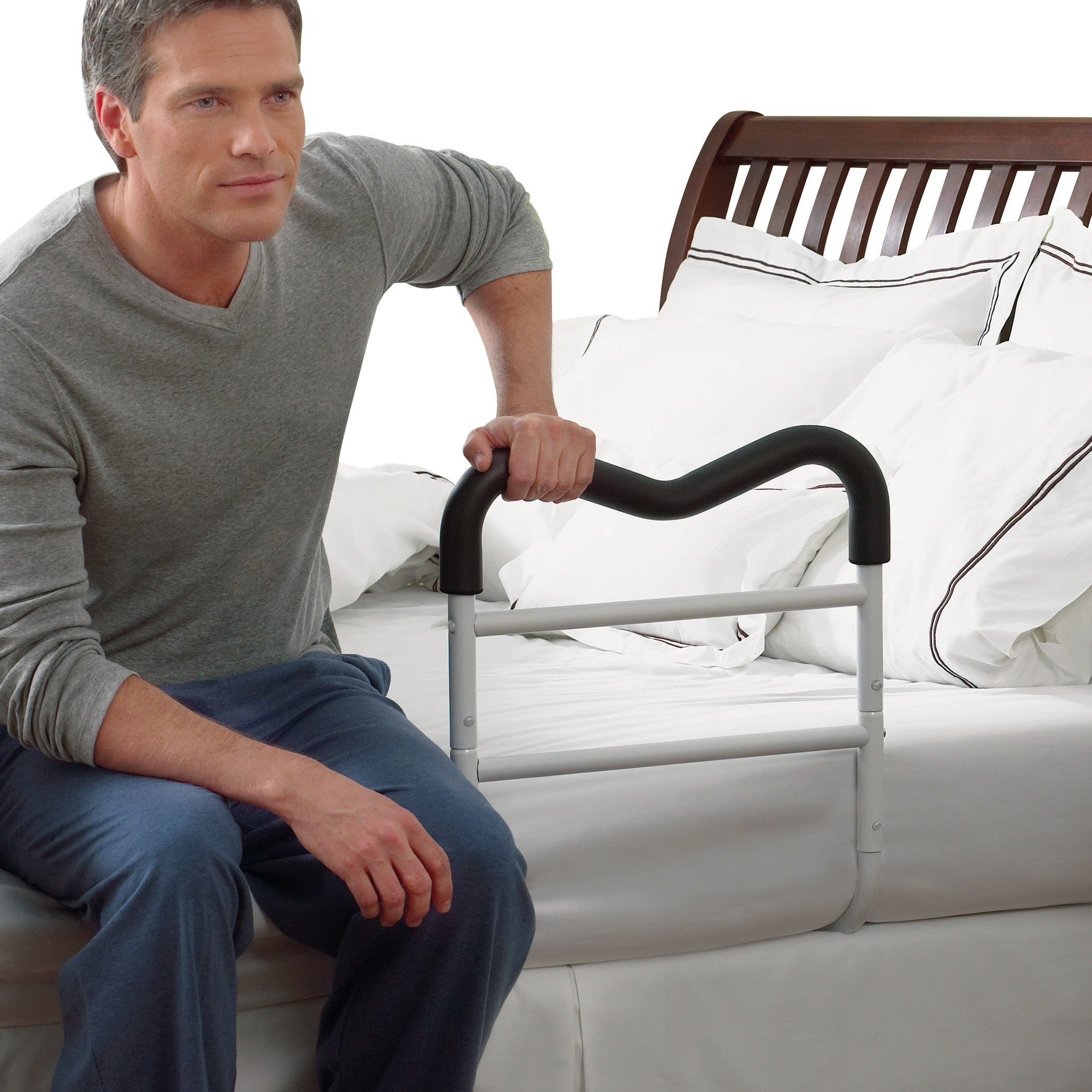 A person uses an M-Rail Assistive Bed Rail, attached to a neatly made bed, for support. The rail is white with a black top handle.