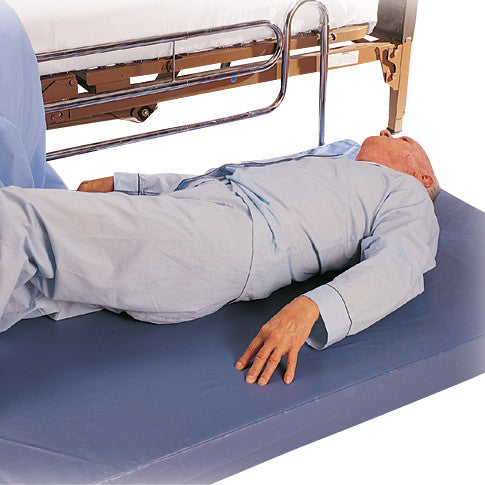 An elderly person lies on a SkiL-Care EasyClean Mat next to a hospital bed, wearing light blue pajamas.