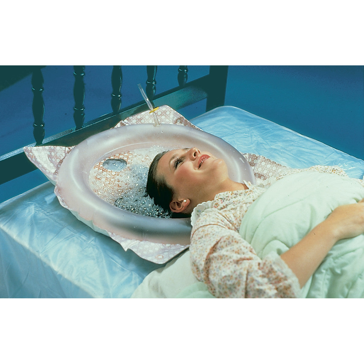 A woman is lying on a bed using a Shampoo Tray to wash her hair. The inflatable tray holds water and soap around her head.