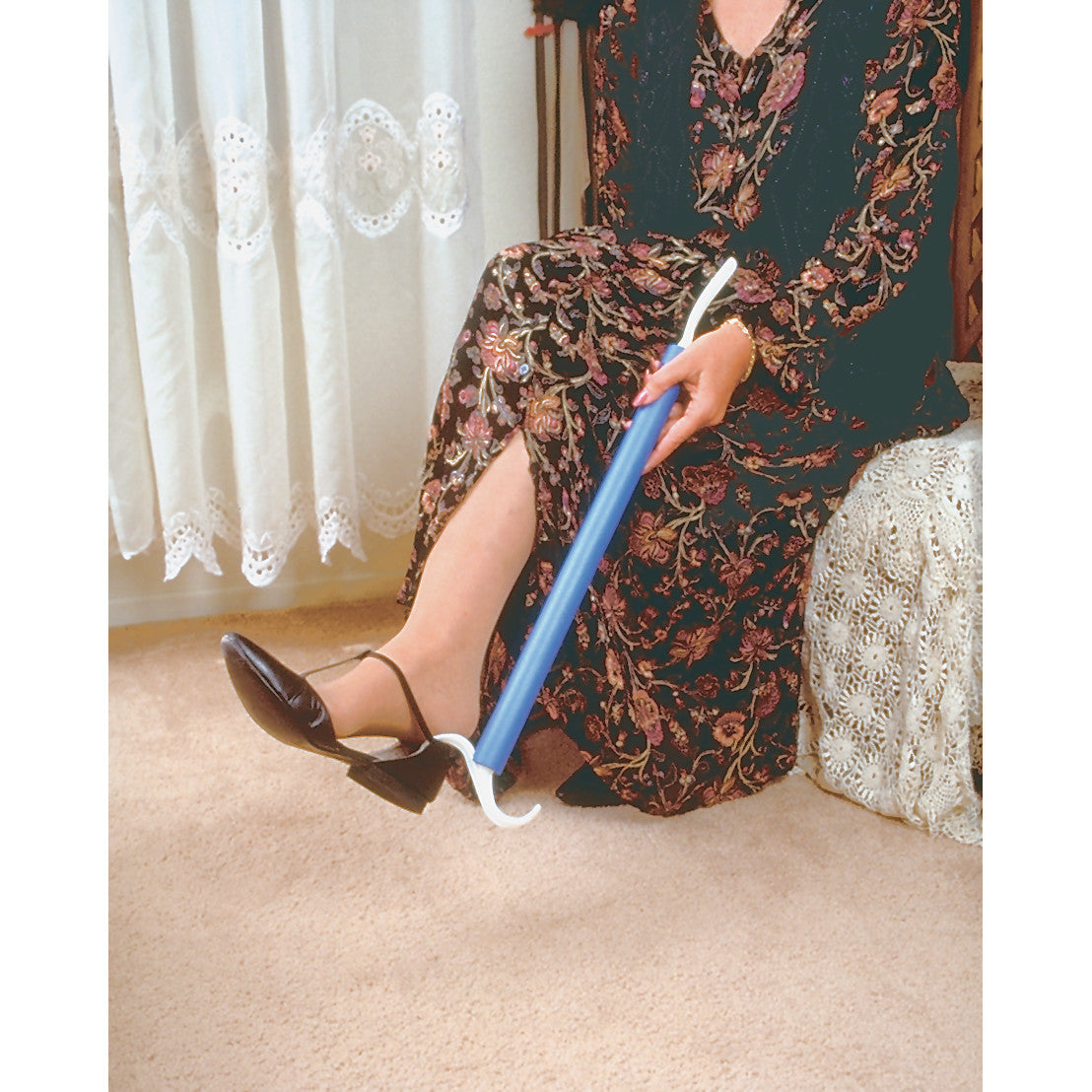 A person uses the Dress EZ assistive tool to adjust a black shoe while sitting, wearing a floral dress in a carpeted room.