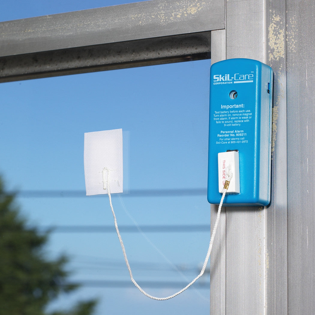 SkiL-Care Window Alarm attached to a window, featuring a blue device with a white pull cord connected to a sensor pad.