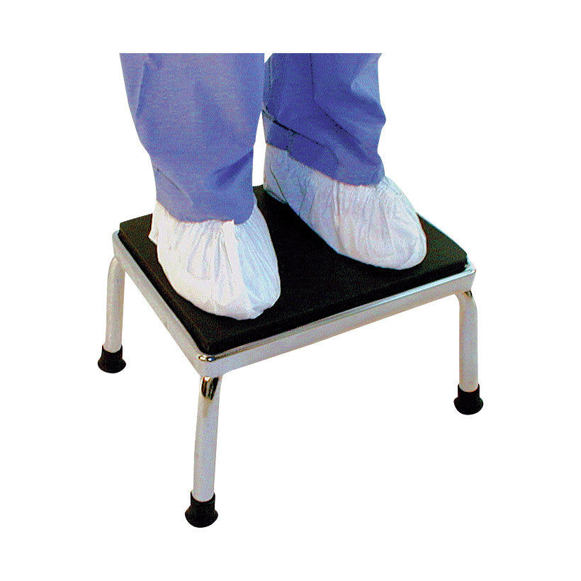 Person standing on an AliMed Soft Step Stool, wearing blue pants and white shoe covers. The stool has a black top and metal legs.