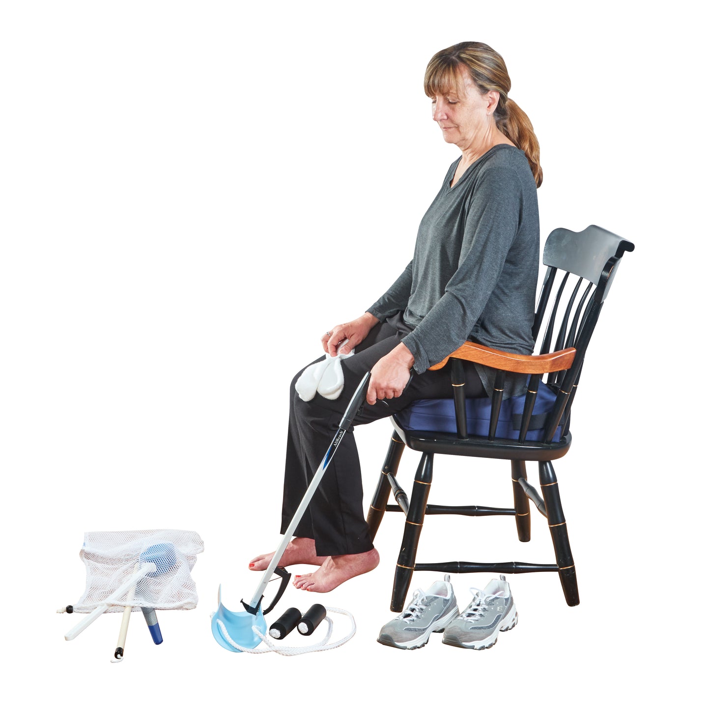 A woman sits on a chair using the AliMed Short ADL Deluxe Hip Kit with Extra-Wide Sock-Aid. Nearby are tools and sneakers.