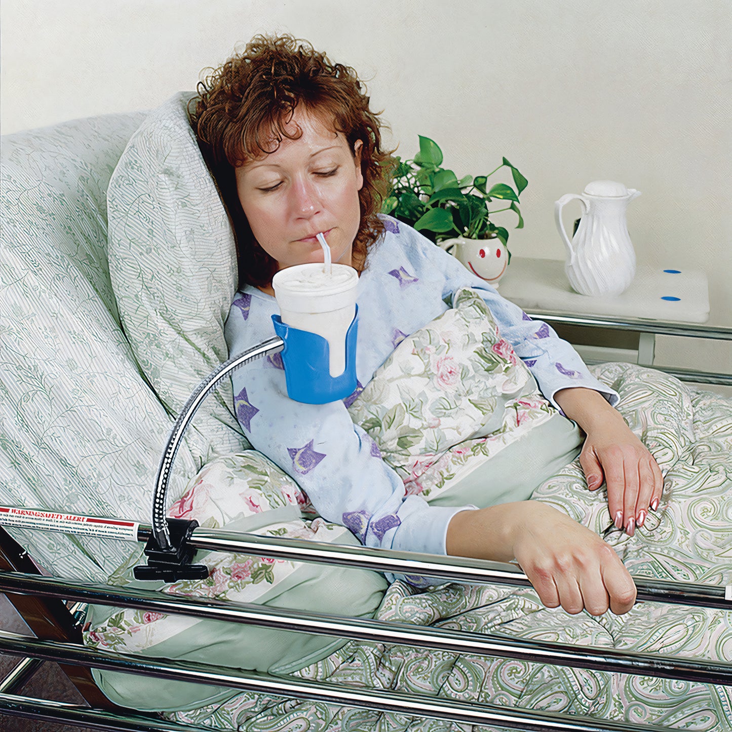 A woman in bed uses a straw to drink from a cup held by a flexible Bedside Beverage Holder attached to the bed railing.