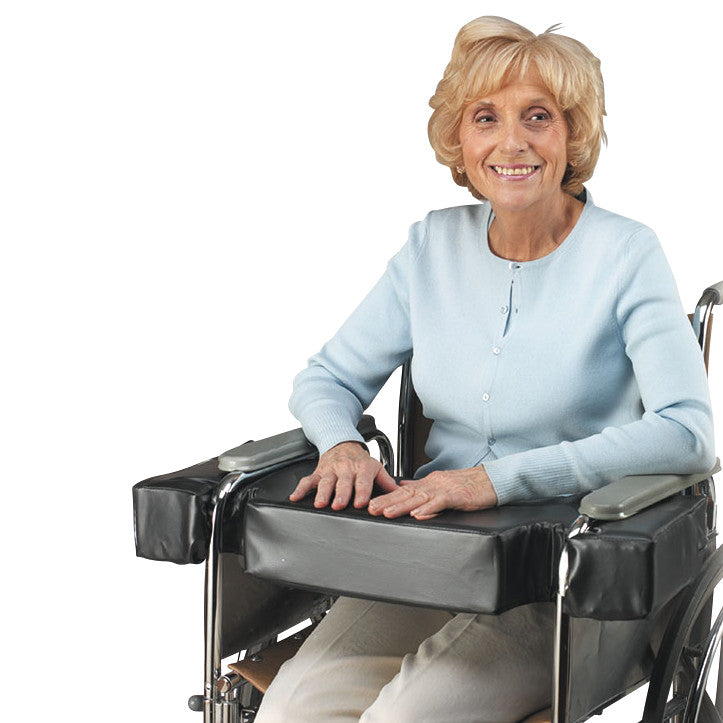 A person sits in a wheelchair with a SkiL-Care LapTop Cushion on their lap. The cushion is black and fits across the chair's arms.