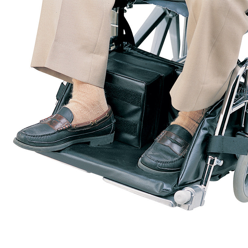 A person in a wheelchair uses a SkiL-Care Foot Cradle, which supports their feet. They wear brown shoes and beige socks.