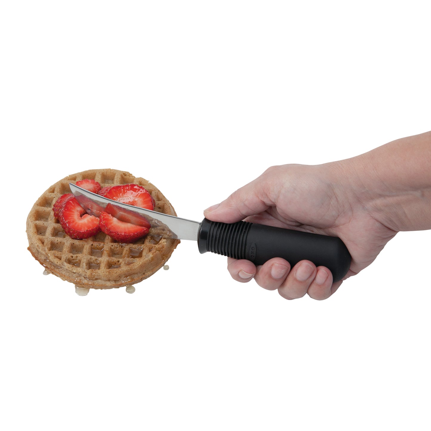 A hand holds a Good Grips Standard Utensil knife, cutting a waffle topped with sliced strawberries.
