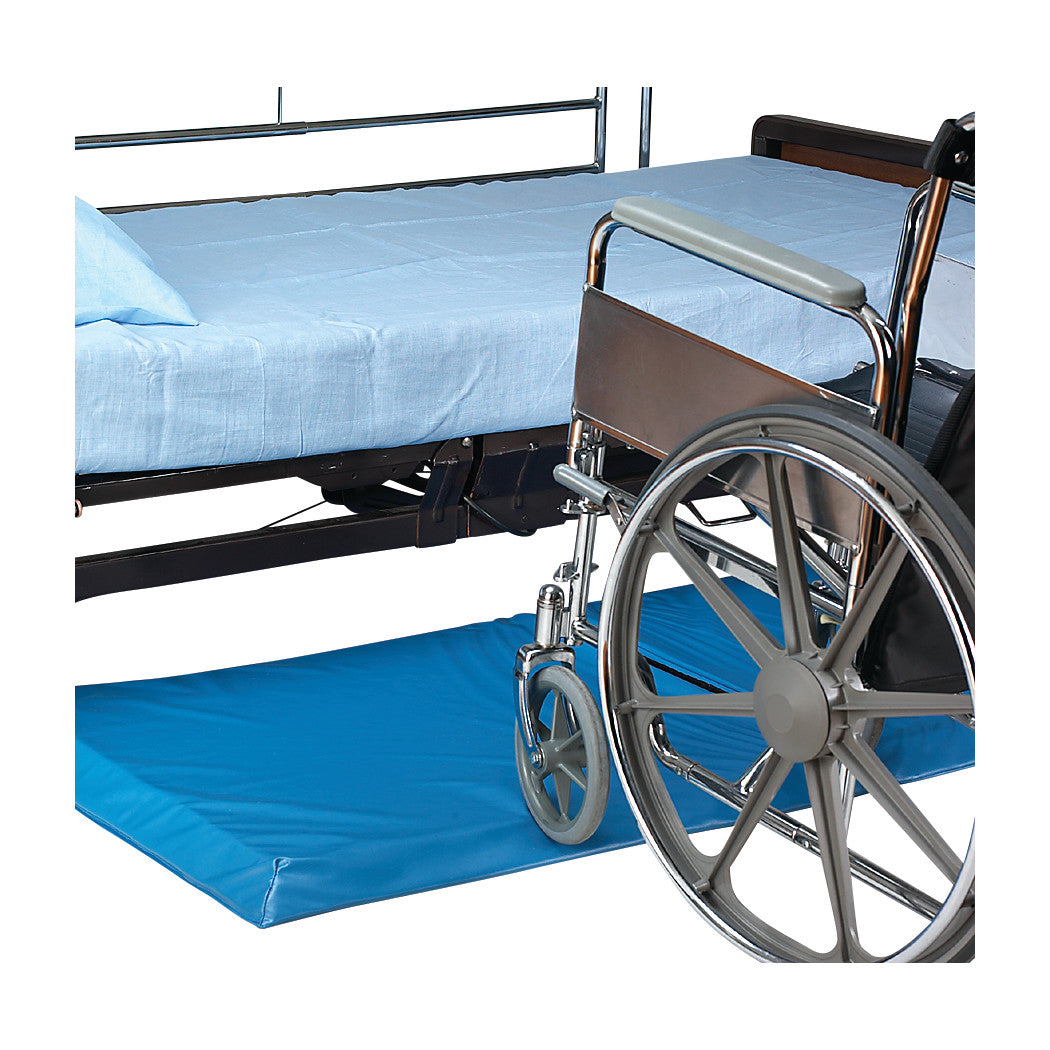 A wheelchair is positioned beside a bed with blue sheets over a SkiL-Care Bi-Fold Roll-On Bedside Mat, providing safety and comfort.