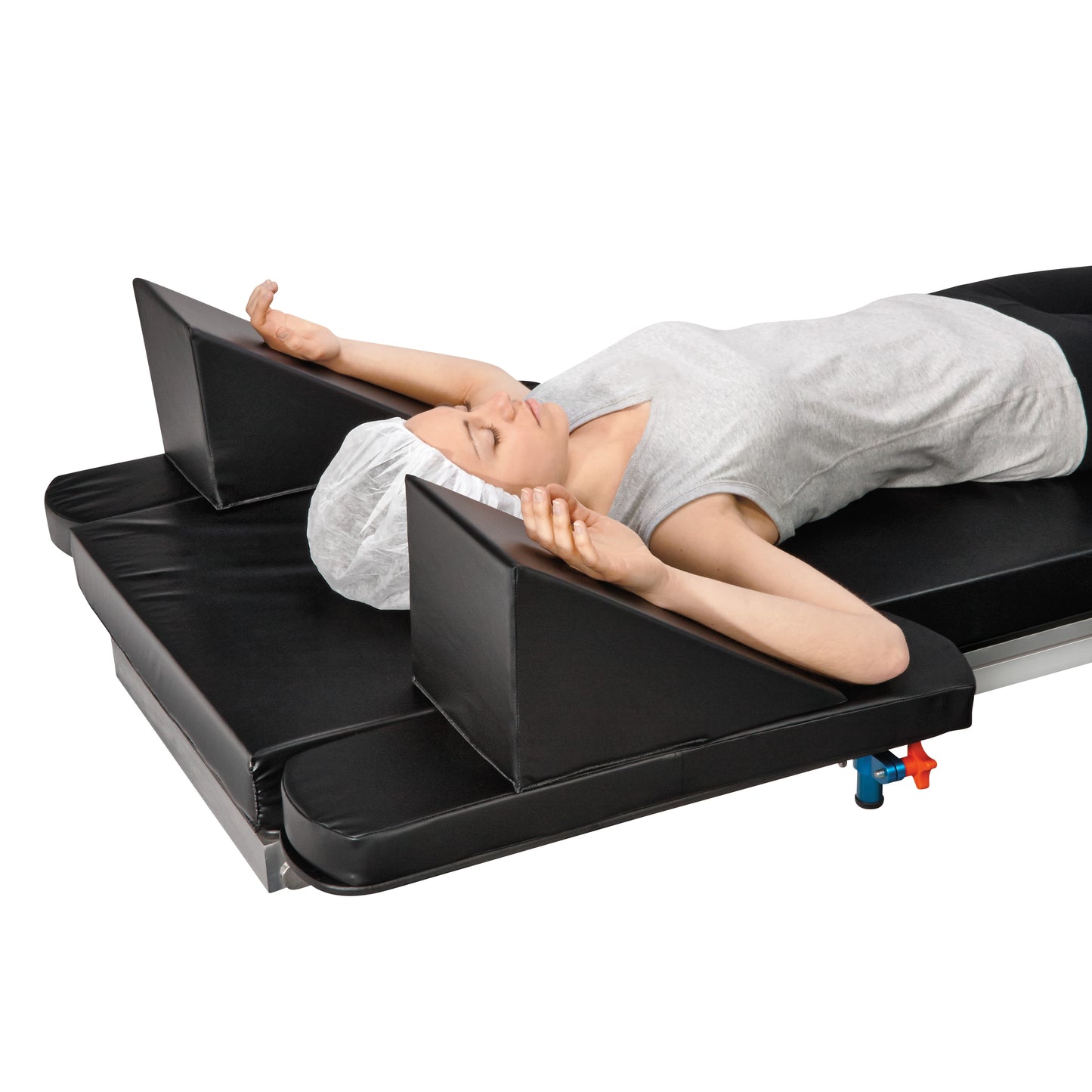 A person lies on their back with arms raised, supported by an AliMed Cardiac Wedge Set on a medical bed, wearing a hair cover.