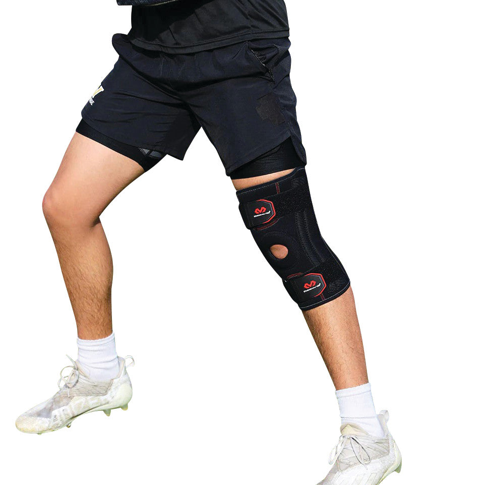 A person wearing a knee brace called the McDavid Patella Knee Support while posing in athletic shorts and white cleats.