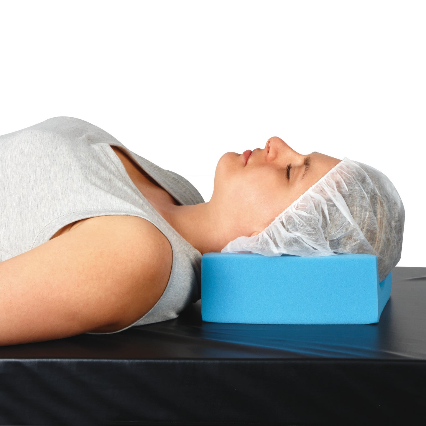 A woman reclines on the AliMed Head/Heel Rest Positioner, a blue foam block, wearing a gray top and hair net, on a black surface.