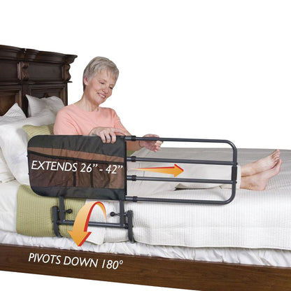 An older adult sits up in bed using the Stander EZ Adjust Bed Rail. The rail extends from 26" to 42" and pivots down 180 degrees.
