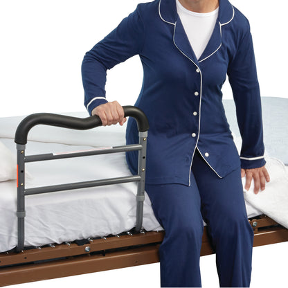 A person in blue pajamas uses the AliMed AliRail Bed Rail, attached to the side of a bed, for support while sitting up.
