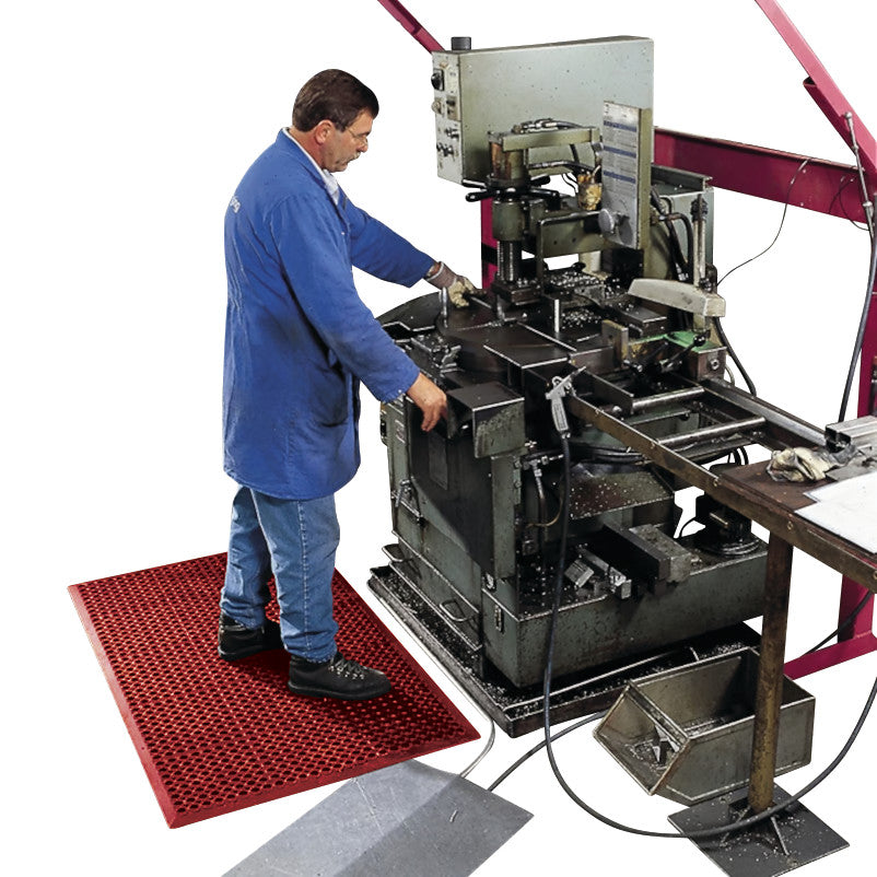 A person in a blue coat operates machinery while standing on a Sanitop Drainage Anti-Fatigue Mat in an industrial setting.