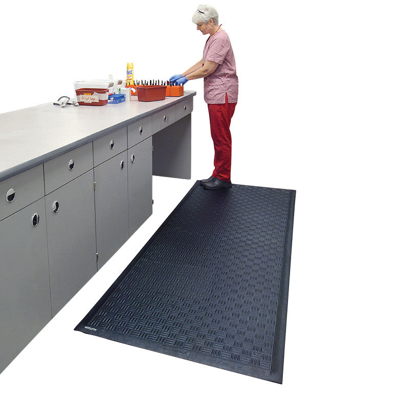 A person stands on a Cushion Station Anti-Fatigue Mat near a counter, organizing items. The mat is black with a textured surface.