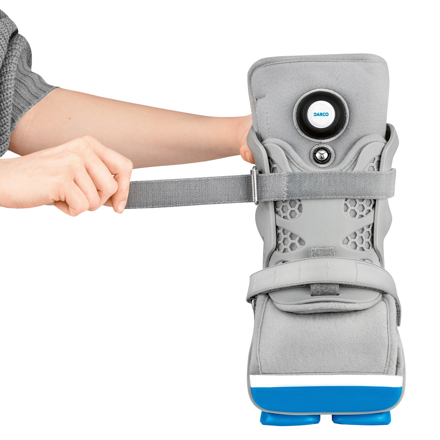 A person is adjusting the strap of a DARCO Body Armor PFA Walker, which is a gray orthopedic boot with blue accents.