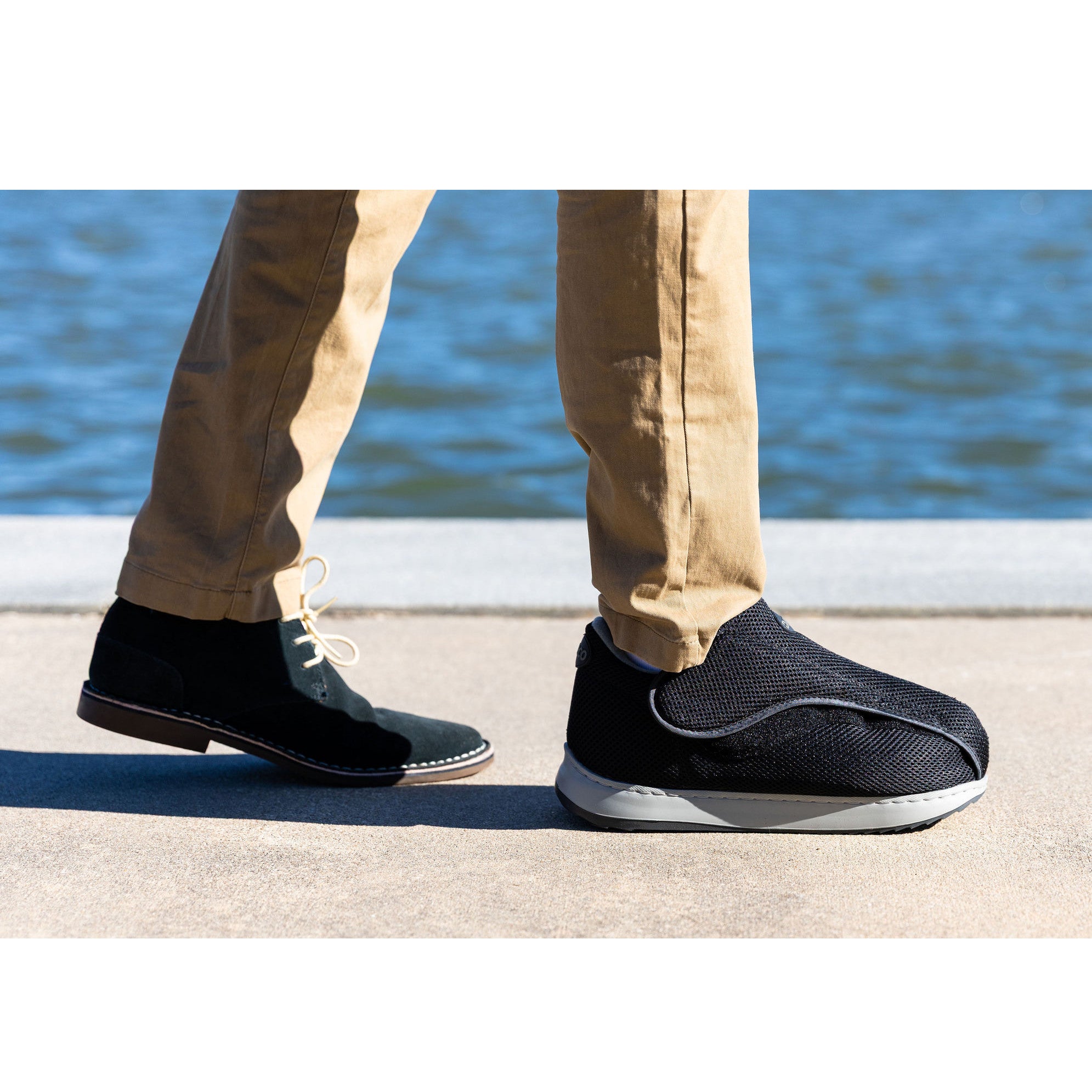 A person wearing tan pants, one black shoe, and a DARCO AllRound Shoe walking by a body of water on a sunny day.