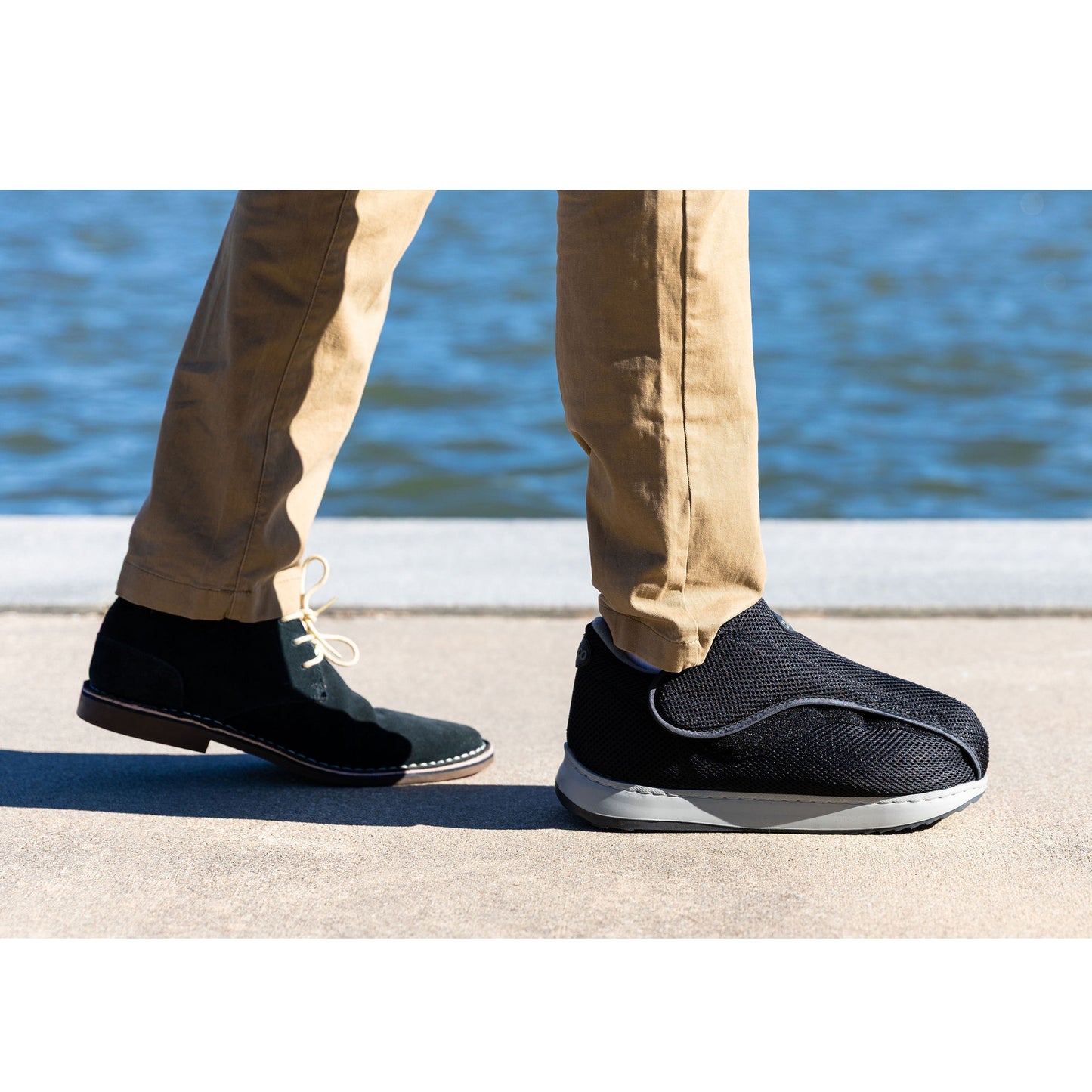 A person wearing tan pants, one black shoe, and a DARCO AllRound Shoe walking by a body of water on a sunny day.