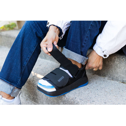 A person adjusts the strap of a DARCO DUO Pro Wound Care Shoe, sitting on concrete steps, wearing rolled-up jeans and a white jacket.