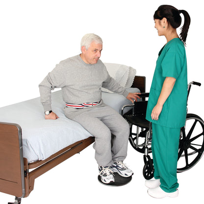 A man sits on a bed with his feet on the SafetySure Pivot Disc, assisted by a caregiver near a wheelchair.