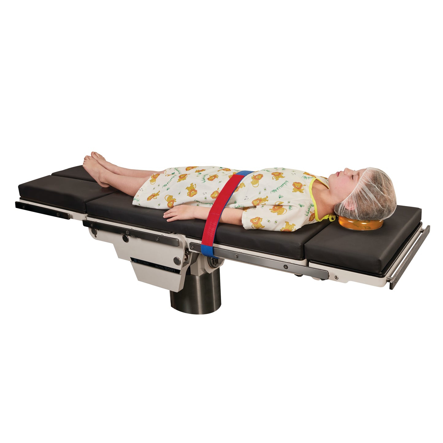 A child lies on a medical table wearing a hair cover and gown, secured with an AliMed Universal Strap, One-Piece, One Closure, No Side Rail.