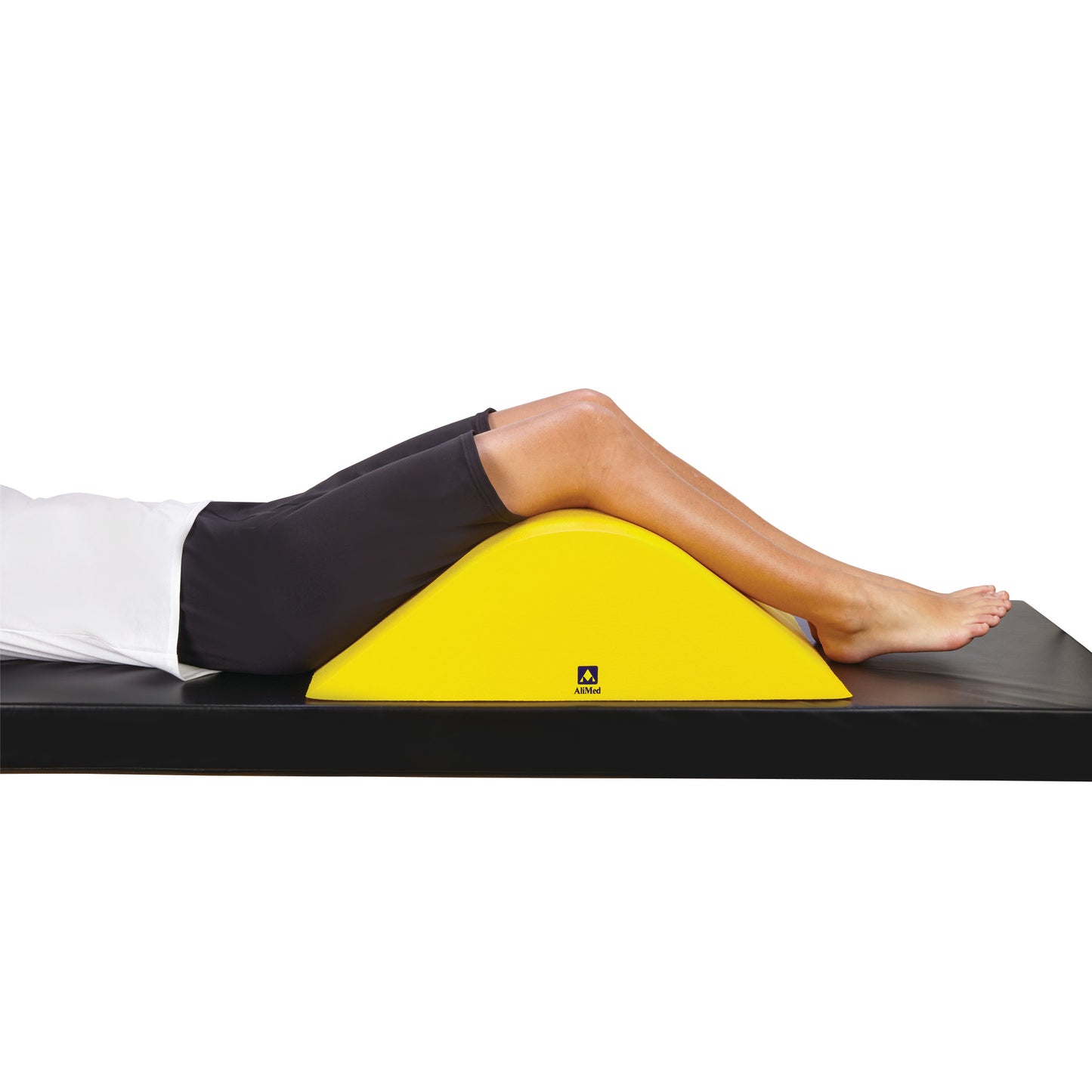 A person rests their legs on an AliMed Protecta-Coat Deluxe Knee Bolster, a yellow, half-cylinder cushion on a black mat.