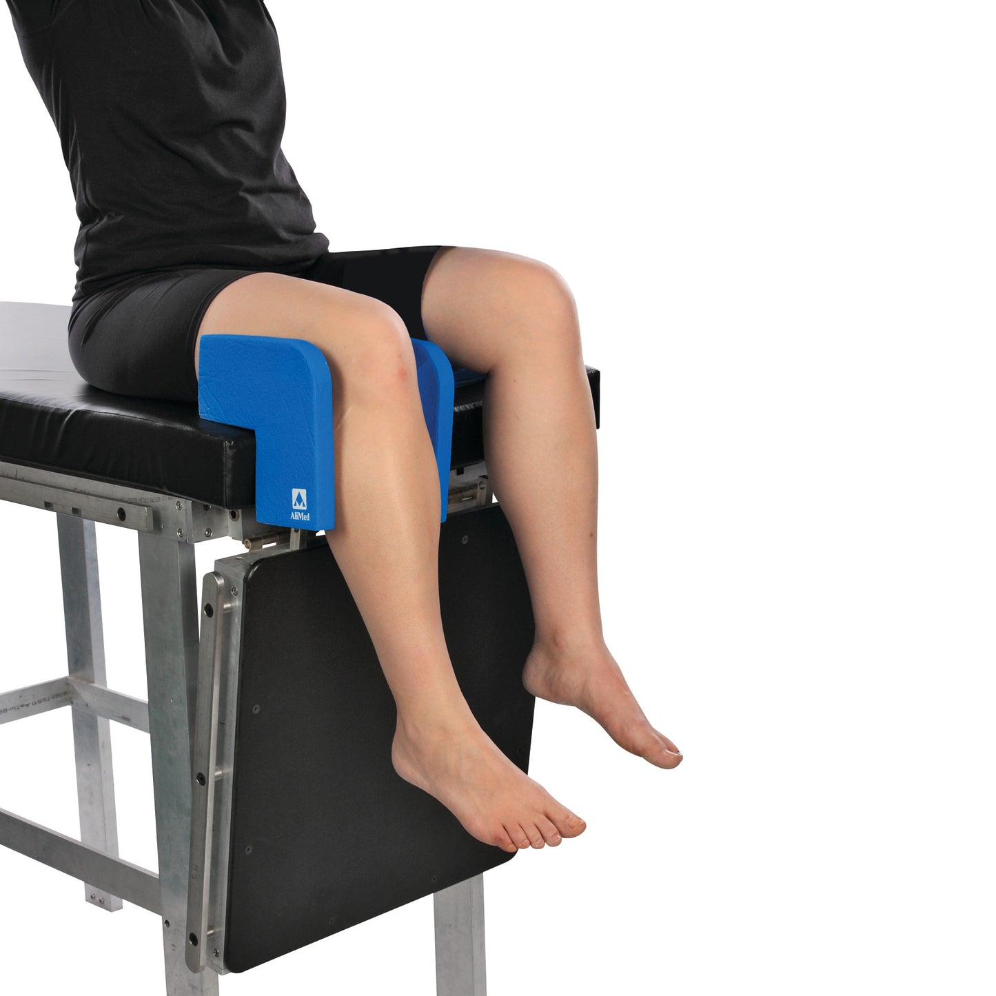 A person sits on a table with legs supported by the AliMed Vinyl-Coated Well-Leg Holder, a blue padded device for stabilization.