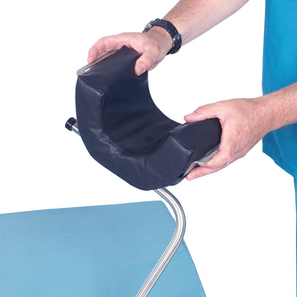A person holds the AliMed Standard Universal Positioner, a cushioned, U-shaped support with a metal frame, above a blue surface.