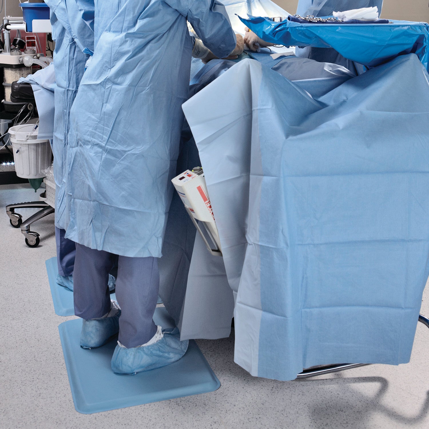Medical staff wearing blue scrubs stand on GelPro Medical Mats during a procedure, surrounded by surgical drapes and equipment.