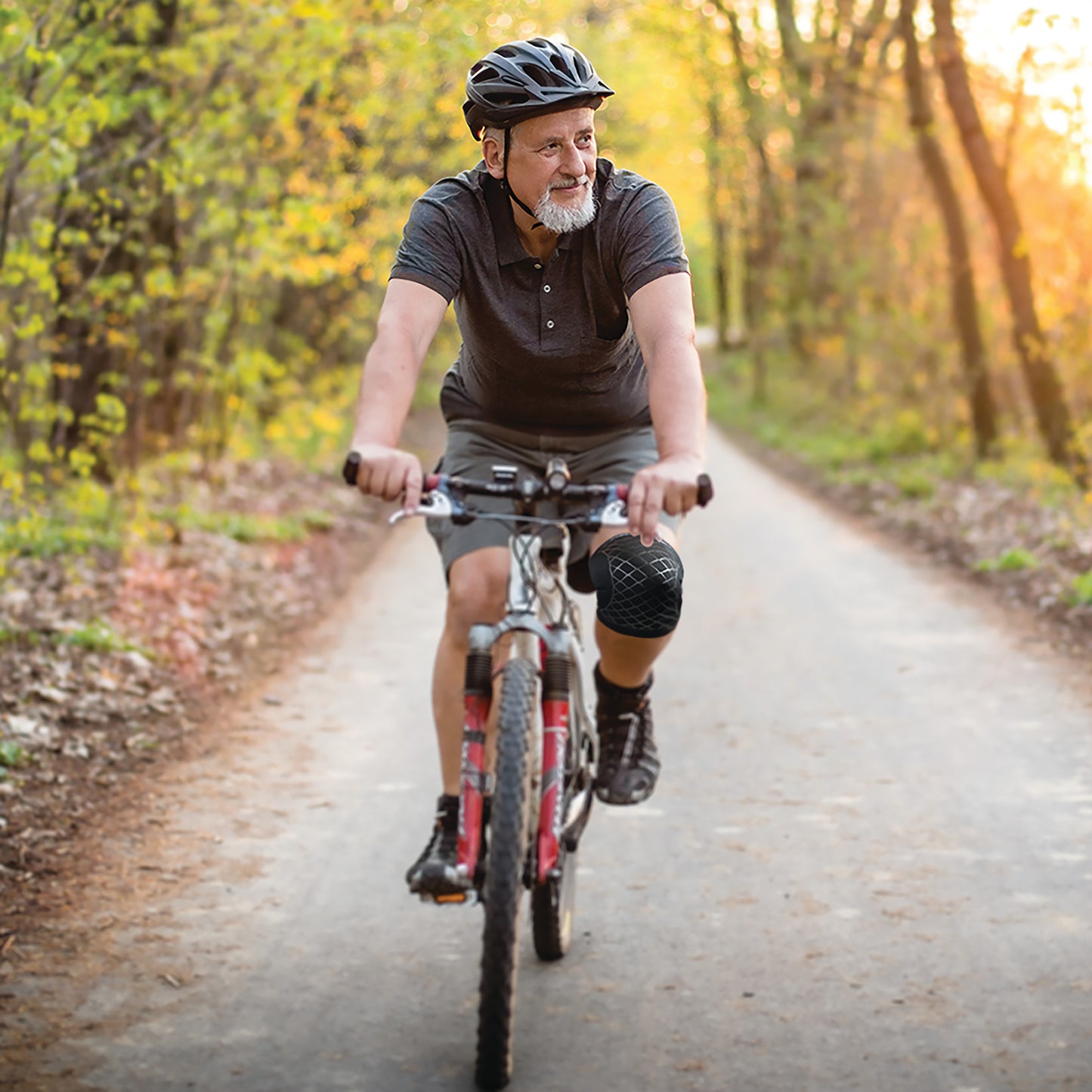 An older man rides a bike on a forest path, wearing a Brownmed Spark Kinetic Knee Sleeve and a black helmet.