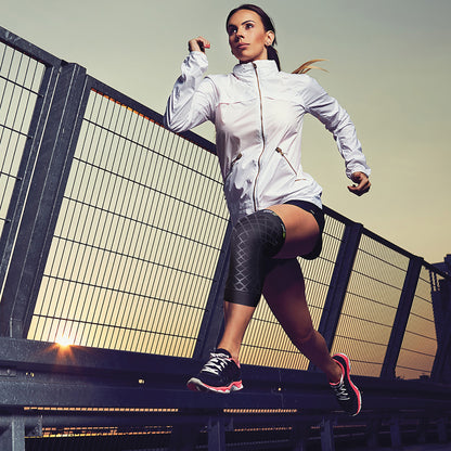 A person is running while wearing a Brownmed Spark Kinetic Knee Sleeve on their left knee, along with athletic gear and shoes.