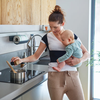 A woman cooks while holding a baby, wearing a Femi Posture support wrap, in a modern kitchen with wooden cabinets.