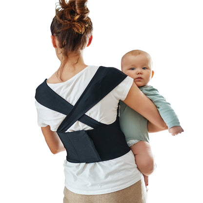 A woman wears a Femi Posture back support while holding a baby. The support is black with straps crossing in the back.