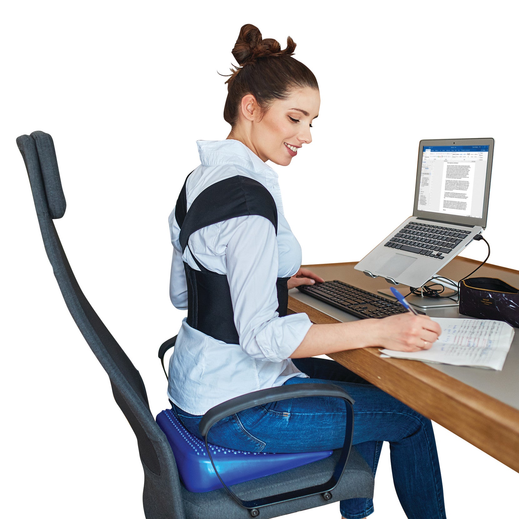 A woman uses a laptop at a desk while wearing the Femi Posture brace. She is sitting on a cushion with a supportive chair back.