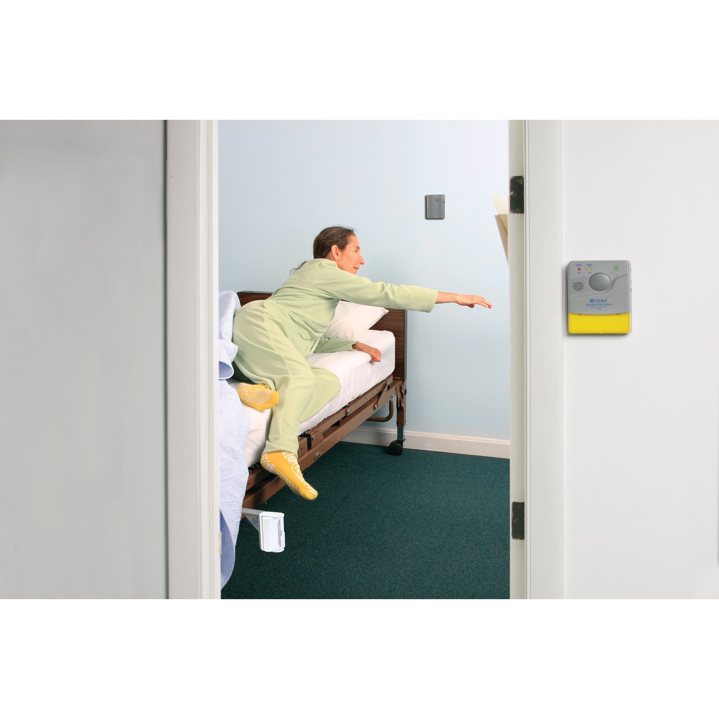 A person reaches from a bed, activating the AliMed Motion Detection Local Alarm with Bed Rail Clamp, mounted on a wall nearby.