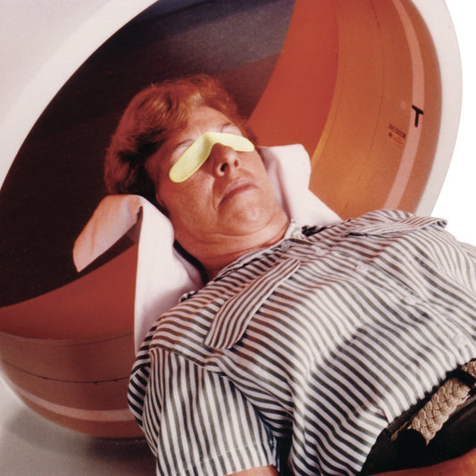 A person lies in a CT scanner wearing an AttenuRad CT Eye Shield with a striped shirt and head supported by pillows.