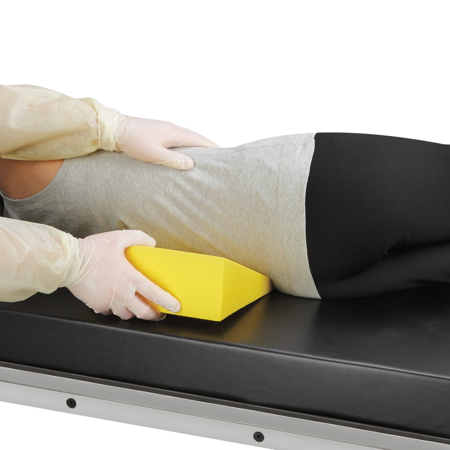 A person wearing gloves positions an AliMed Protecta-Coat 15 Degree Wedge Positioner under a patient's back on a black medical table.