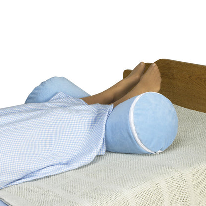 A person rests on a bed with feet elevated by a SkiL-Care Ultra-Soft Bed Bolster. The bolster is light blue and cylindrical.