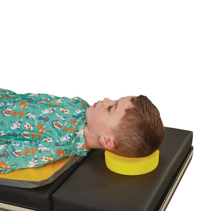 A child rests on a medical bed, head supported by a yellow AliMed Protecta-Coat Head Donut, wearing a teal animal-print gown.